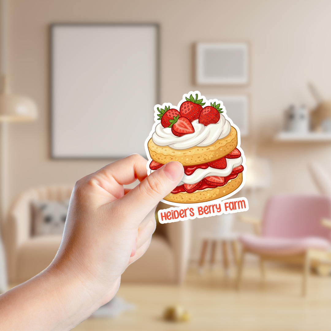 Hand holding a sticker of a strawberry shortcake with 'Heider's Berry Farm' text in a blurred room setting.