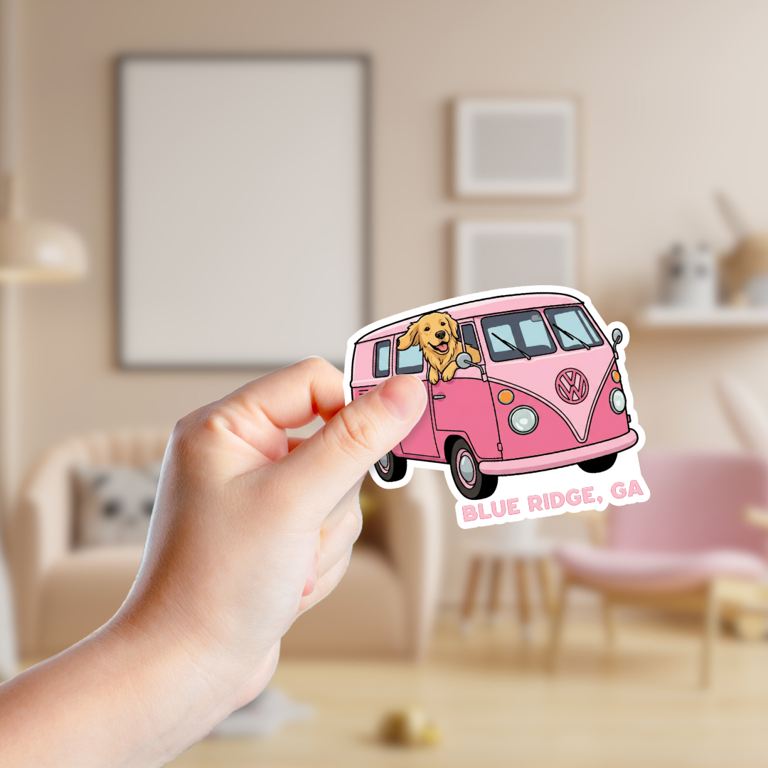 Hand holding a pink Volkswagen van sticker with a dog on a blurred indoor background